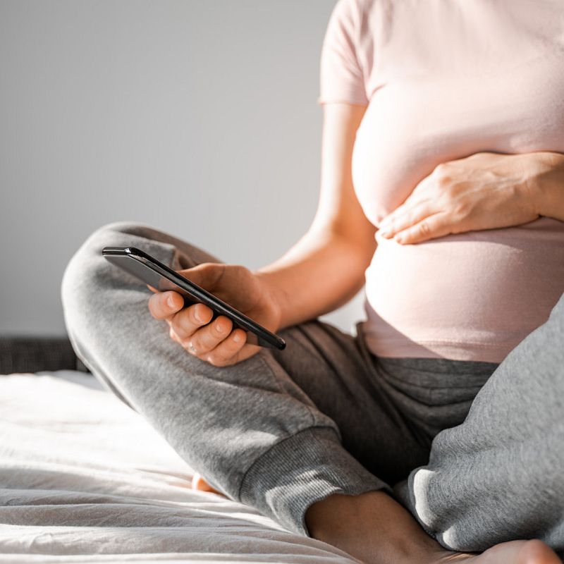 A long-term pregnant woman in the 9th month uses a mobile application on her mobile phone to plan and monitor her pregnancy, preparing to become a mother at home in bed