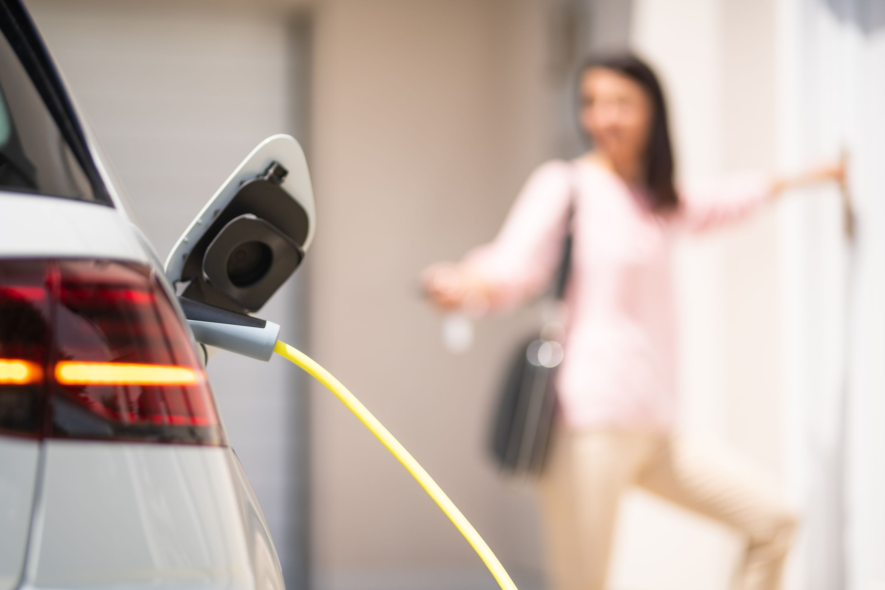 Close up of a electric car charger with female silhouette in the background, entering the home door and locking car