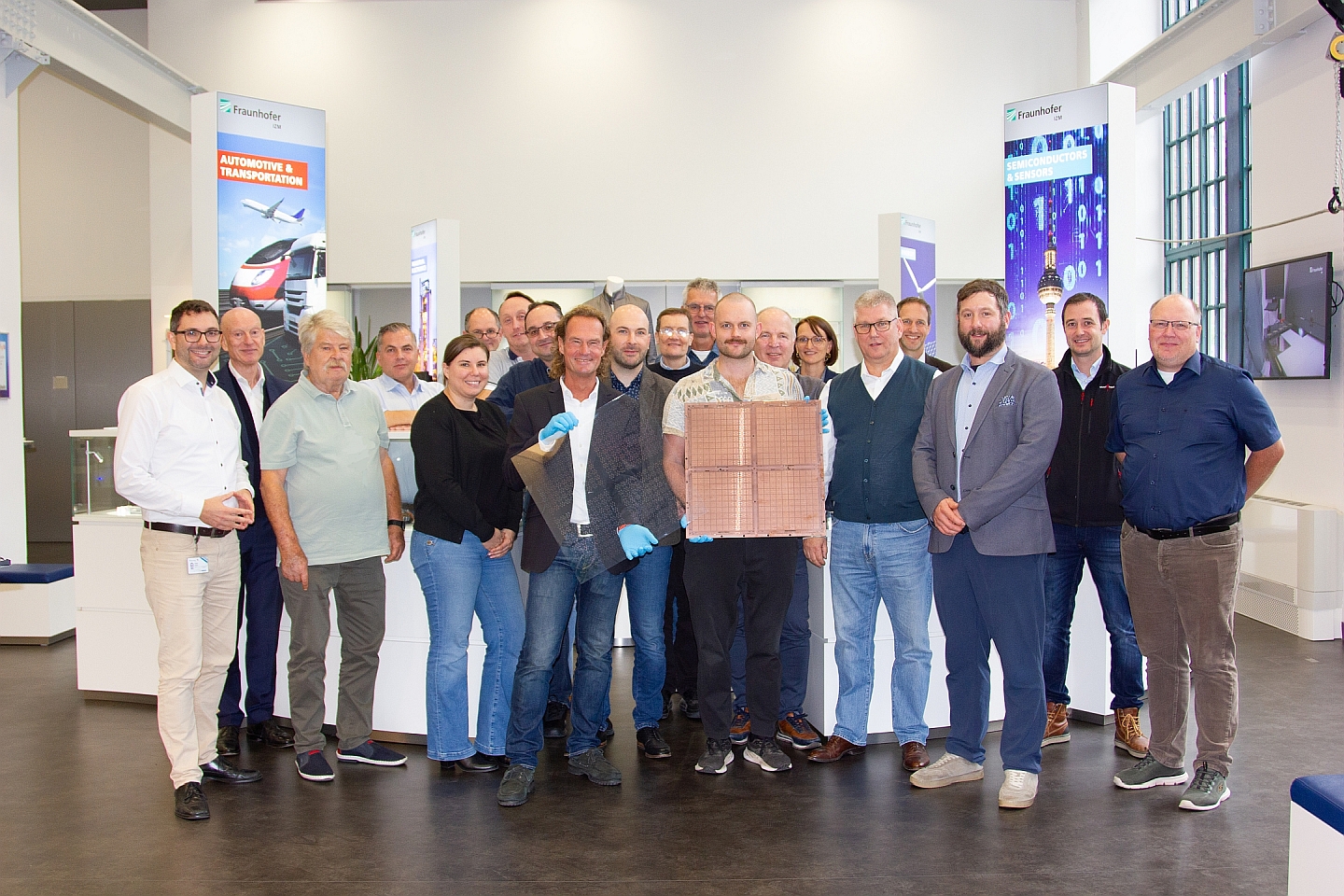 A group of people in a modern exhibition space, posing together. Some individuals are holding glass substrates.