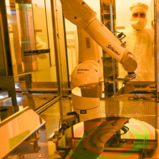 An automated robotic arm machine working on a silicon wafer in a cleanroom environment. A person in protective clothing is visible in the background.